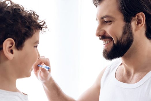 Happy father brushes teeth of young son with toothbrush.